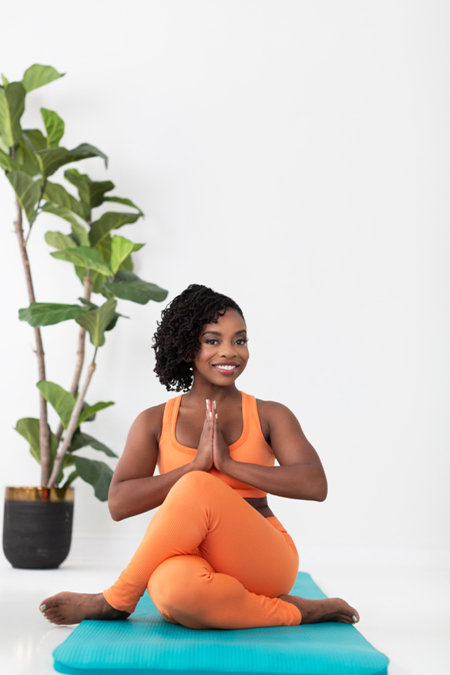 Christina Thompson seated cross-legged on a turquoise yoga mat beside a leafy plant
